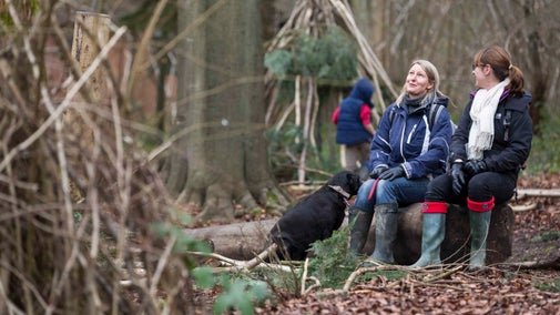 Two women with a black dog sitting on a tree stump and chatting in woodland at Basildon Park, Berkshire.